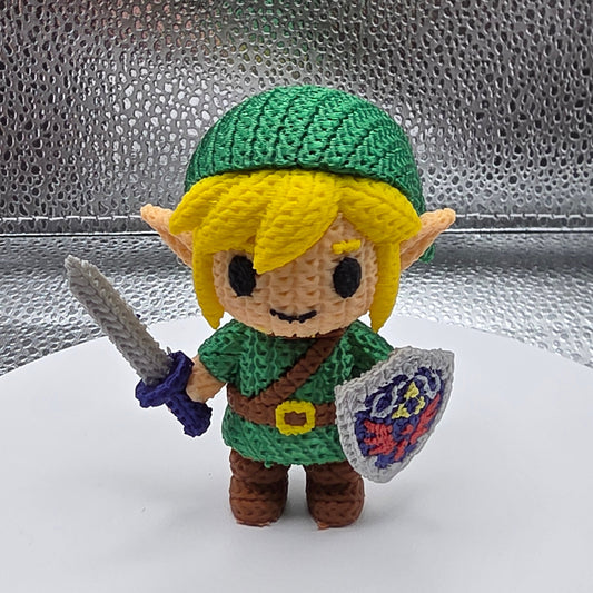 Crocheted character with green outfit and yellow hat on a metallic surface